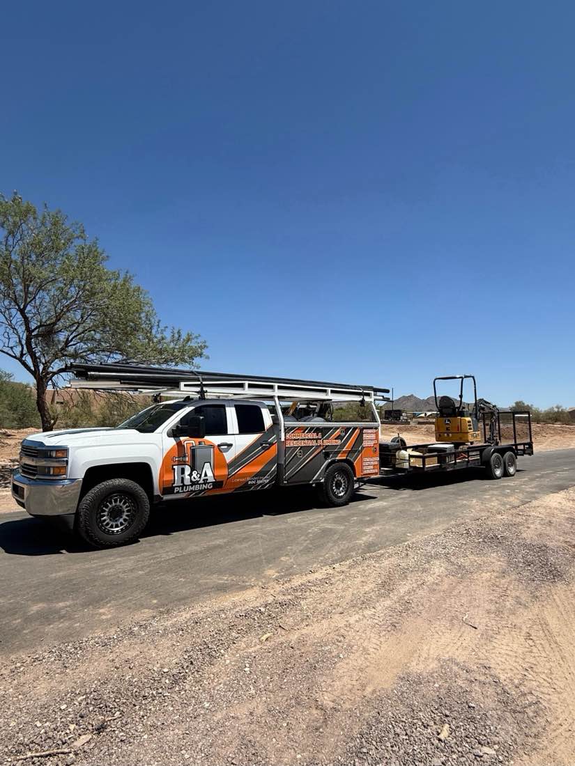 R&A Plumbing service truck towing a trailer with a mini excavator on a desert road.